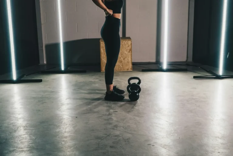 A woman in black workout clothes standing on a gym floor beside a black kettlebell, shown from the neck down