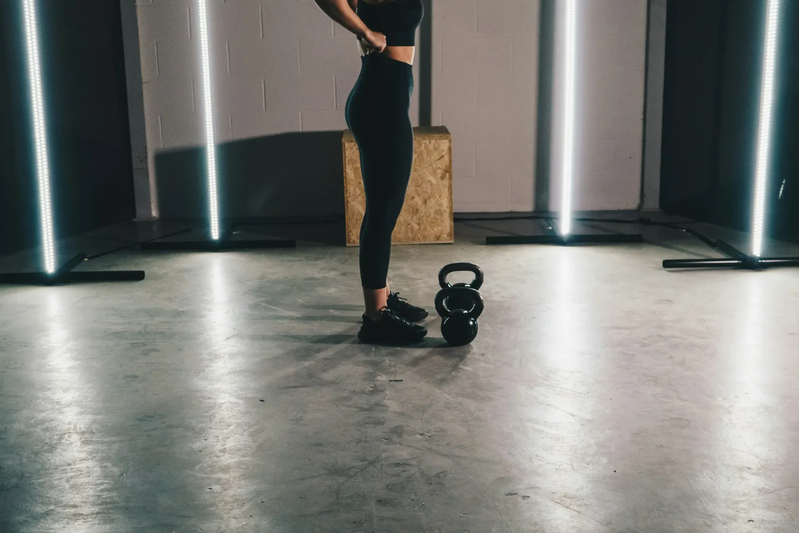 A woman in black workout clothes standing on a gym floor beside a black kettlebell, shown from the neck down