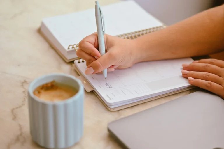 A person writing in a paper planner with a pen, a mug of coffee on the table beside it