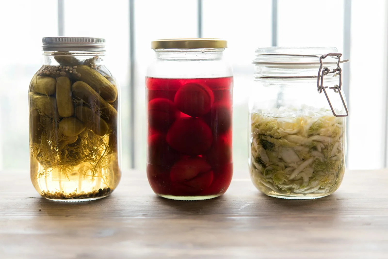 Three glass jars of homemade fermented vegetables, pickled cucumbers, beets and sauerkraut, on a table by a bright window