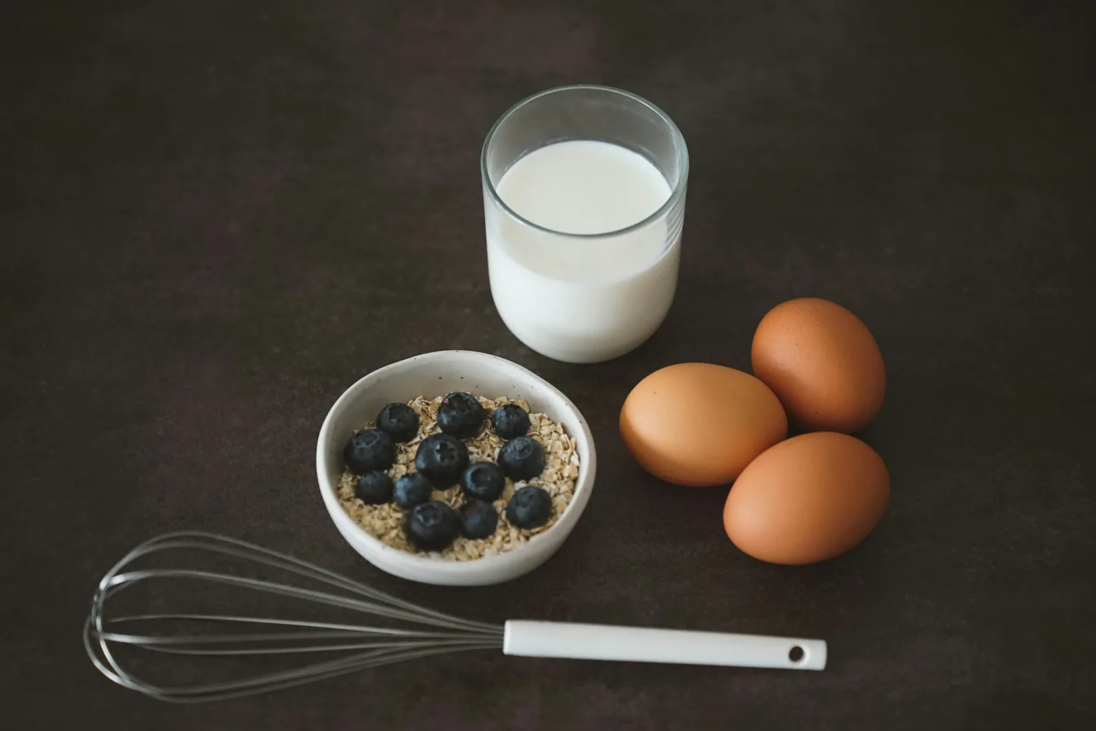 A glass of milk, three brown eggs and a small bowl of oats topped with blueberries on a dark table
