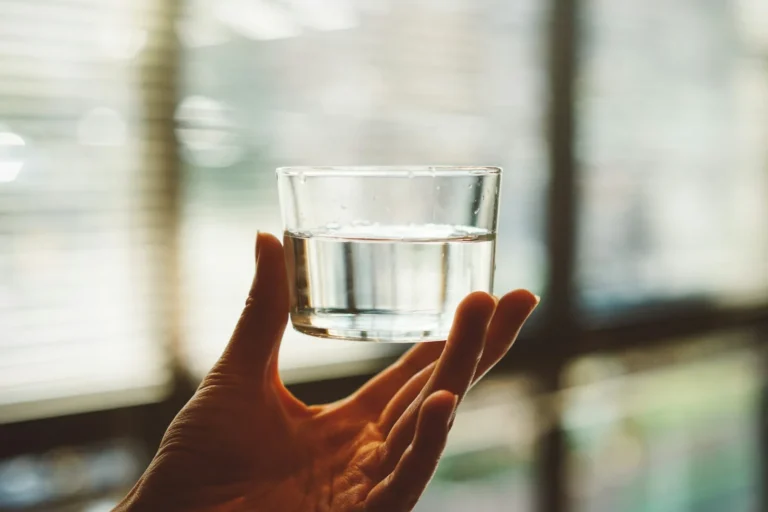 A hand holding a small clear glass of water in front of a window with blinds