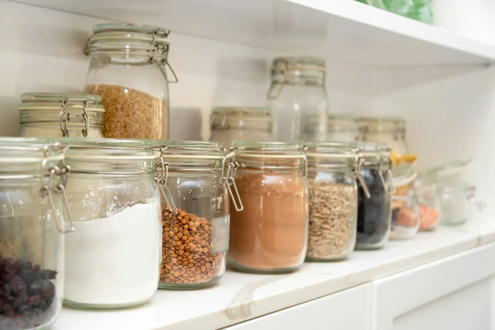 A row of glass storage jars on a white shelf holding lentils, grains, seeds and dried fruit