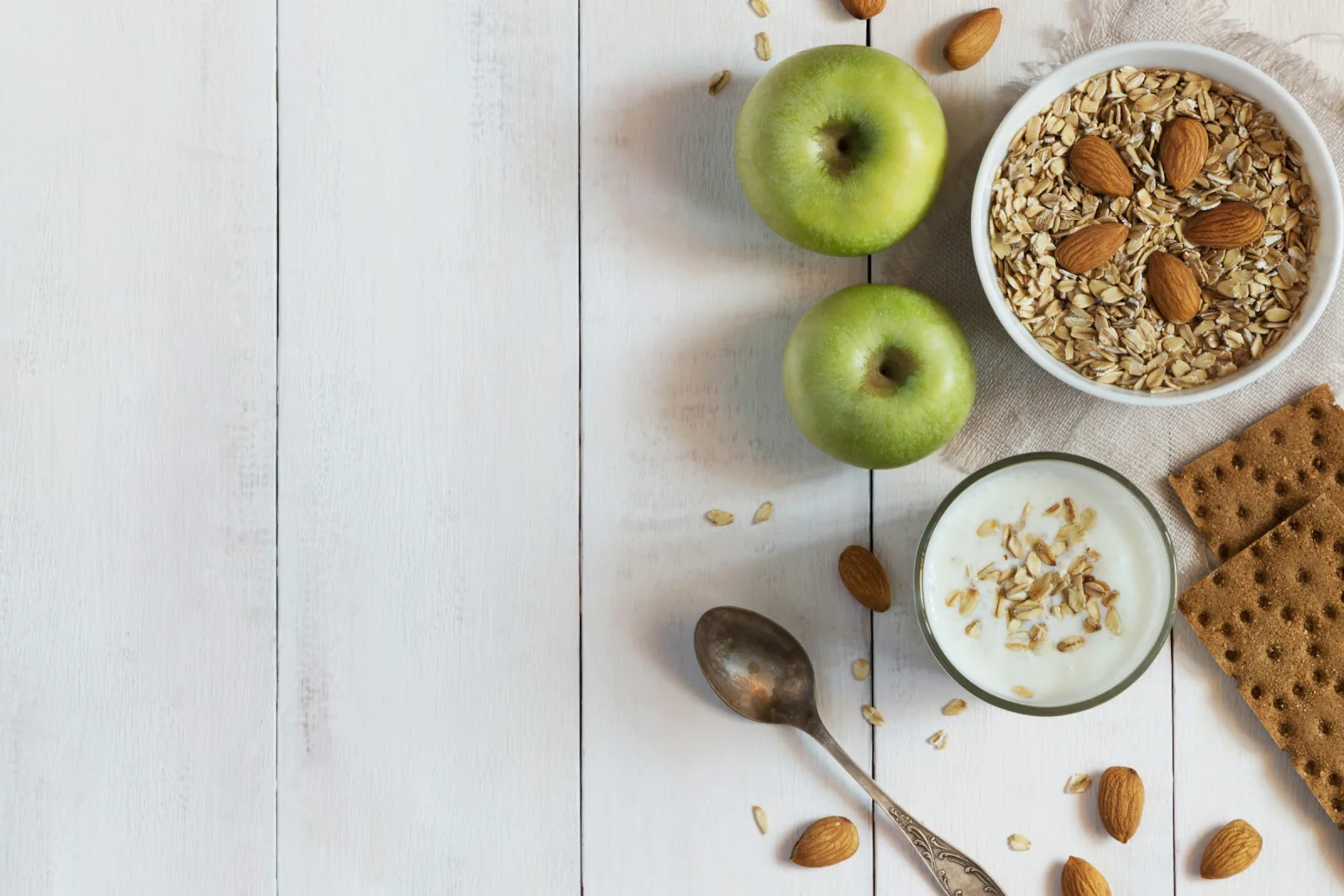 A white table with a bowl of rolled oats and almonds, a glass of yogurt topped with oats, two green apples and crispbread